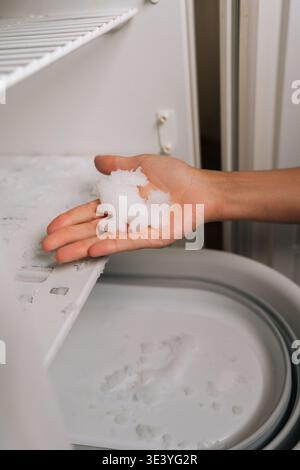 Male hand holding sizeable ice block from freezer defrosting, melting fragments collecting in underlying containers during kitchen cleaning Stock Photo