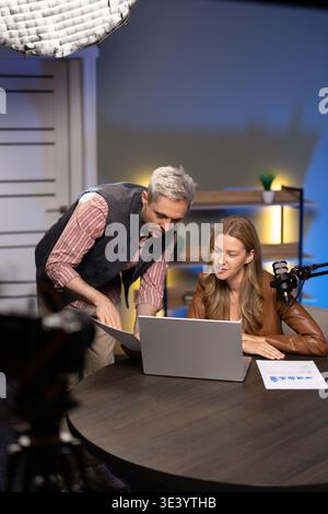 Podcasters reviewing printed analytics reports, collaborating using laptop in studio Stock Photo