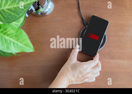 Top view of woman's hand charging smartphone with wireless charger on wooden desk. Modern technology, portable fast charger concept. Stock Photo