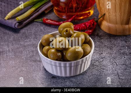 Close-up still life of green olives in a white ramekin. A rustic scene with red chili peppers, scallions, and oil on a dark, textured surface. Appetiz Stock Photo