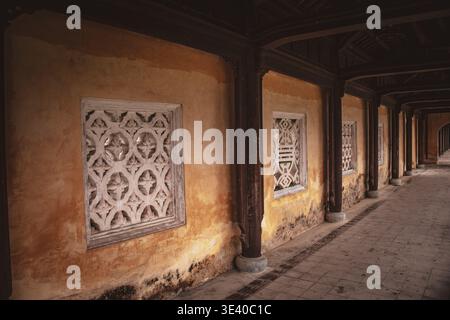 Yellow corridor wall with carved stone lattice windows and dark timber columns receding into distance, Hue, Vietnam. Stock Photo