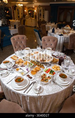 Round table covered with silver velvet tablecloth, laden with diverse dishes: soups, appetizers, mains, desserts, and fruits. Stock Photo