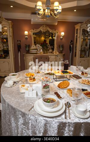 Beautifully set round dining table covered with silver-grey textured tablecloth, filled with appetizers, soups, pastries, fresh fruits and elegant whi Stock Photo