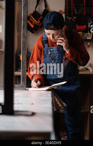 Female carpenter in overalls talking on phone and taking orders in a small craft workshop. copy space Stock Photo