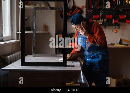 Female carpenter in overalls talking on phone and taking orders in a small craft workshop. copy space Stock Photo
