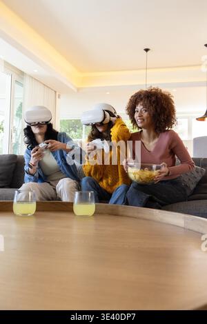Diverse female friends wearing white VR headsets holding controllers, sharing chips on sofa at home Stock Photo