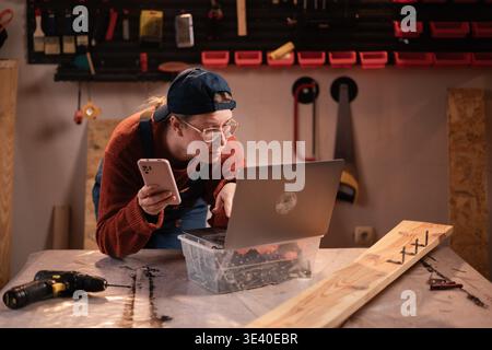 Carpenter Working In Woodwork Workshop Talking On Mobile Phone And Using Laptop Stock Photo