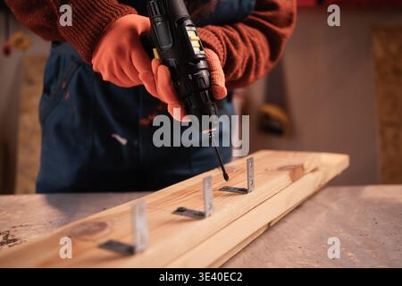 Female Carpenter drilling hole into timber block working in her workshop. Woman doing furniture assembling in studio. Woodworking expert using electri Stock Photo