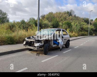 Wrecked and dismantled silver hatchback car abandoned on the side of a suburban road with missing parts and a shattered windshield. Stock Photo