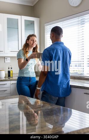 Diverse couple standing by countertop in kitchen, woman holding ceramic mug near horizontal blinds Stock Photo