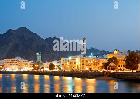 People walk along the waterfront in Muscat, Oman during sunset. The sky shows colors as lights turn on in the buildings. Stock Photo