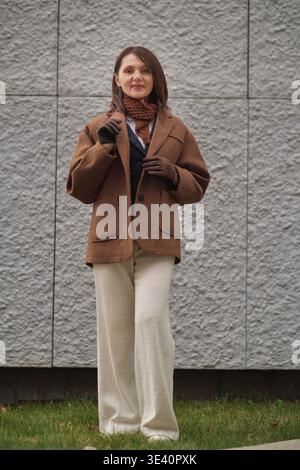 A woman in autumn attire stands on grass near a modern wall. The scene represents harmony, refined taste, and the aesthetic of quiet confidence. Stock Photo