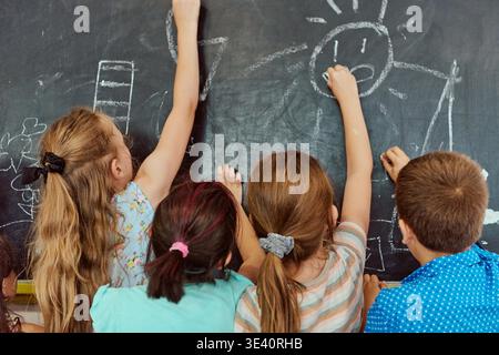 Four elementary students draw with chalk on a classroom blackboard, collaborating and expressing creativity while learning through play. Back-view captures joyful interaction, hands-on art activity and cooperative learning. || Model released Stock Photo