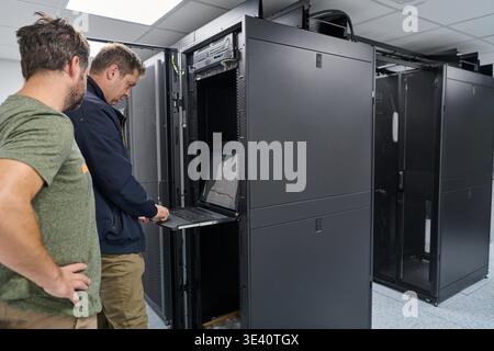 Two IT technicians inspect and configure servers in a modern data center, using a rack-mounted laptop for diagnostics and teamwork while maintaining network hardware and infrastructure in a secure server room. || Model released Stock Photo