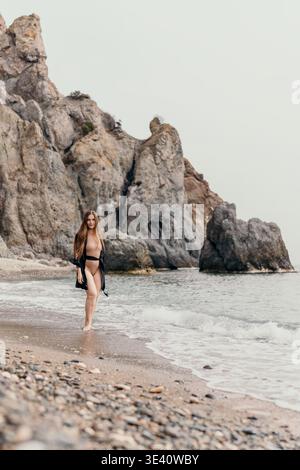 Woman beach cliffs walking on sandy shore with rocky mountains in background under overcast sky Stock Photo