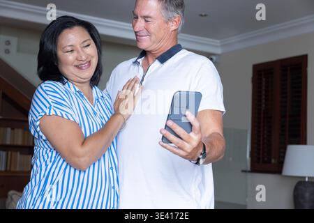 Mature couple Asian woman leaning on man in casual clothes, taking smartphone selfie in living room Stock Photo