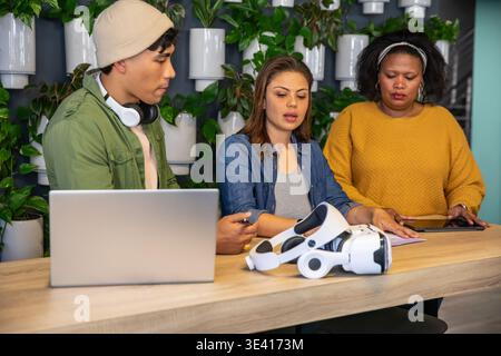 Diverse coworkers collaborating at counter in coworking space with VR headset, laptop, plant wall Stock Photo