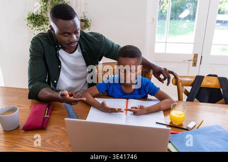 African American father and school-age son leaning at table, father guiding homework with laptop Stock Photo