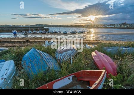 Upturned dinghies and kayaks rest in grassy dunes beside Elie harbor as fishing boats sit on the mud at low tide, the sun bursting through clouds and Stock Photo