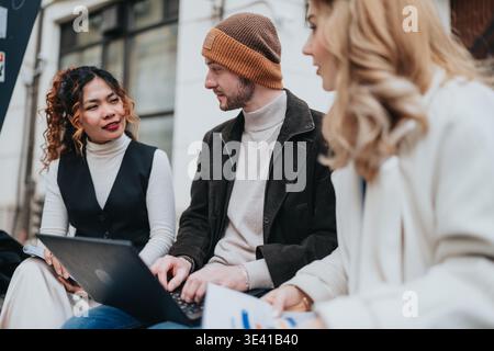 Three young professionals collaborating outdoors on a laptop during an informal business meeting Stock Photo