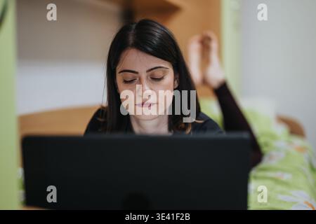 Young woman working on a laptop while lying on her bed at home, focused on the screen Stock Photo