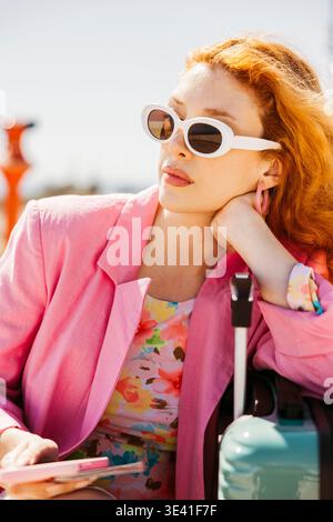Woman with red hair wearing sunglasses and a pink blazer resting on her luggage Stock Photo