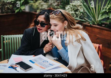 Two female colleagues in a cafe reviewing business documents and sharing a phone call Stock Photo