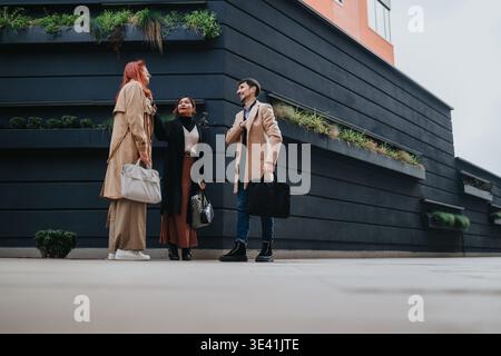 Three colleagues having a casual business meeting outside a modern office building in autumn coats Stock Photo