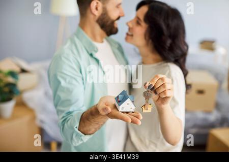 Smiling couple with keys and tiny house model, unpacking boxes in new home Stock Photo