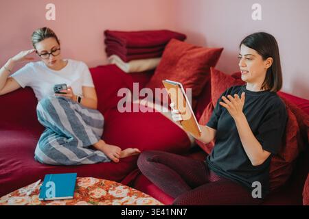 Two female friends having a video call on a tablet while relaxing on a red couch Stock Photo