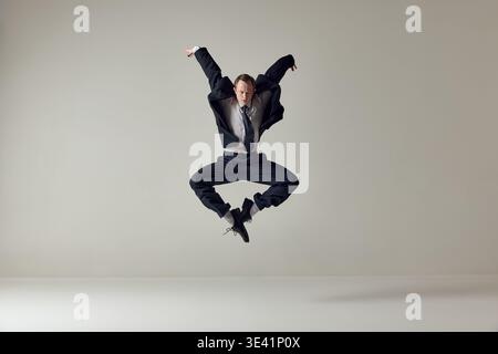 Businessman jumping with arms raised in expressive motion on neutral background. Stock Photo