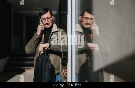 Businessman checking watch while talking on phone outside office with reflective glass wall Stock Photo