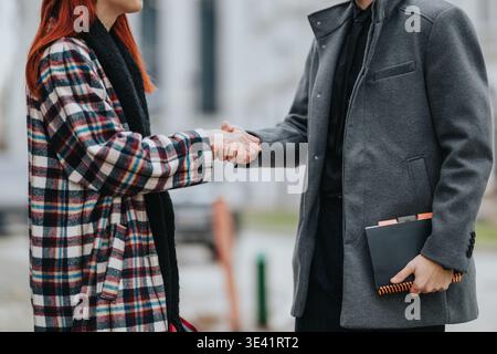 Business partners shake hands outdoors after a meeting, woman in plaid coat and man with notebook Stock Photo