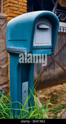 Blue freestanding mailbox with curved top, mounted on post near brick wall and gate. Stock Photo