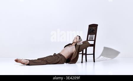 Male dancer lying on floor near chair in relaxed pose in minimal studio Stock Photo