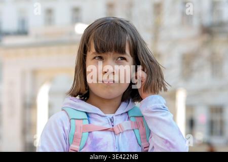 Thoughtful girl about 8 years old with backpack looking aside on city street. Stock Photo