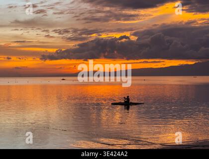 Silhouette of a man on a small raft at sunset on Little Paliton Beach, Siquijor, Philippines, with calm waters reflecting vivid orange skies. Stock Photo