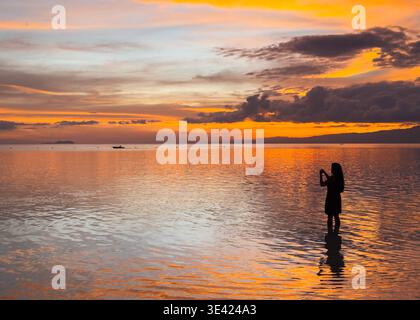 Silhouette of a woman at sunset on Little Paliton Beach, Siquijor, Philippines, with calm sea reflecting vibrant orange and golden skies. Stock Photo