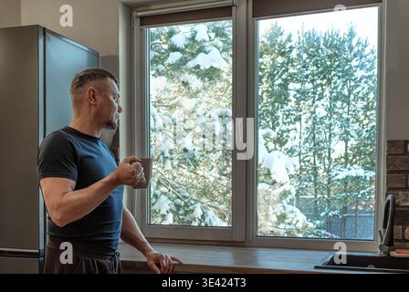 Thoughtful man with mug of hot drink looking out of window to snow scene. Cold winter, living alone, suburban retirement concept. Stock Photo