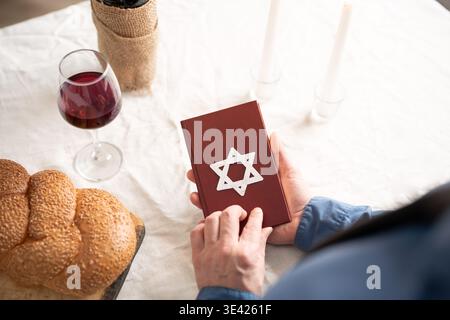 Torah book in hands next to challah bread, kosher wine and burning Shabbat candles on white tablecloth, top view. Shabbat Shalom concept. Reading reli Stock Photo