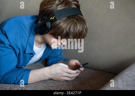 Young man with smartphone and headphones lying on sofa at home.  Cyberbullying, digital overload, video game addiction concept. Stock Photo
