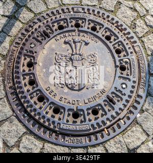 Cast-iron manhole cover bearing the inscription ‘Leipzig City Sewerage System’ and the city coat of arms, set into cobblestones; close-up. Leipzig, Saxony, Germany Stock Photo
