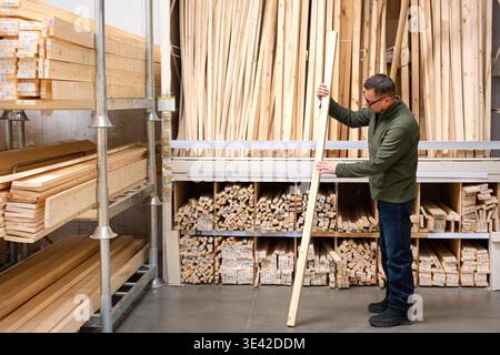 Man selecting timber (wooden bar) for DIY project. Man seller checking quality of lumber board in hardware store or wood warehouse. Stock Photo