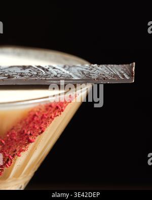Dark chocolate shard rests on cocktail rim. Crisp red flakes adorn creamy drink below. Deep black background enhances visual contrast. Perfect for lux Stock Photo