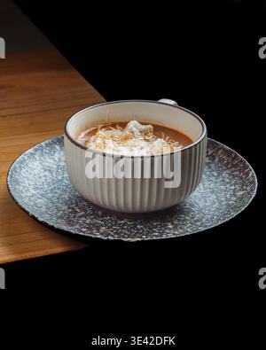 Warm soup sits in a textured ceramic bowl. Crispy garnish floats atop rich, inviting broth. Dark speckled saucer rests on rustic wooden surface. Soft Stock Photo