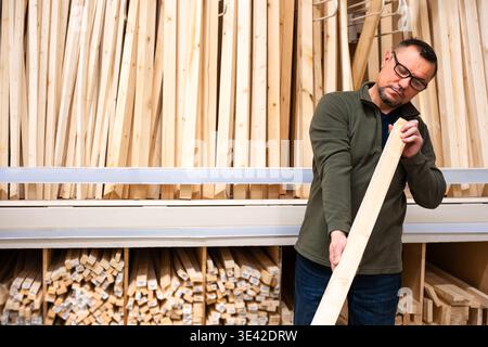 Man selecting timber (wooden bar) for DIY project. Man checking straitness of lumber board in hardware store or wood warehouse. Stock Photo