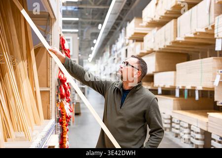 Man selecting timber (wooden lath) for DIY project. Man checking quality of lumber in hardware store or wood warehouse. Stock Photo