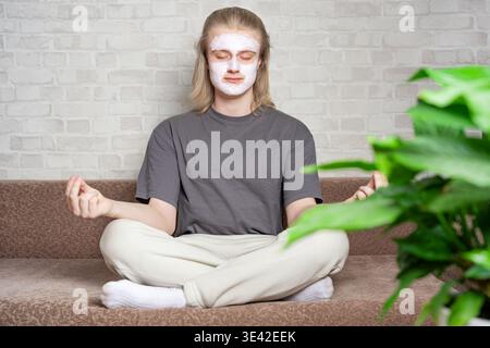 Relaxed young man closing eyes and sitting in metitation pose with cosmetic clay mask on his face. Relaxing, balanced lifestyle, home beauty treatment Stock Photo
