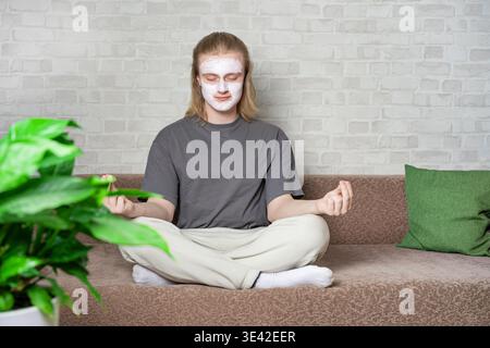 Relaxed young man closing eyes and sitting in metitation pose with cosmetic clay mask on his face. Relaxing, balanced lifestyle, home beauty treatment Stock Photo