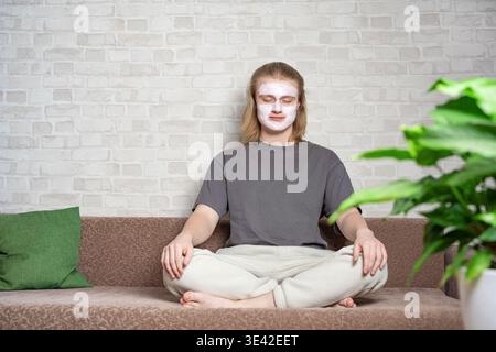 Relaxed young man closing eyes and sitting in metitation pose with cosmetic clay mask on his face. Relaxing, balanced lifestyle, home beauty treatment Stock Photo